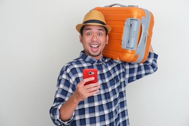 O homem asiático adulto carrega uma bagagem e segura o handphone com expressão alegre