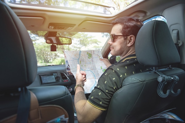 O homem asiático motorista bonito senta-se na mão do carro segura o mapa e vira o corpo para trás com um sorriso e o polegar.