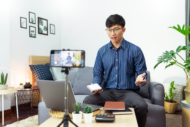 O homem asiático nos fones de ouvido que escreve notas no caderno que olha o curso video do webinar estuda através do estudo em linha da leitura do portátil em casa, conceito do ensino eletrónico.