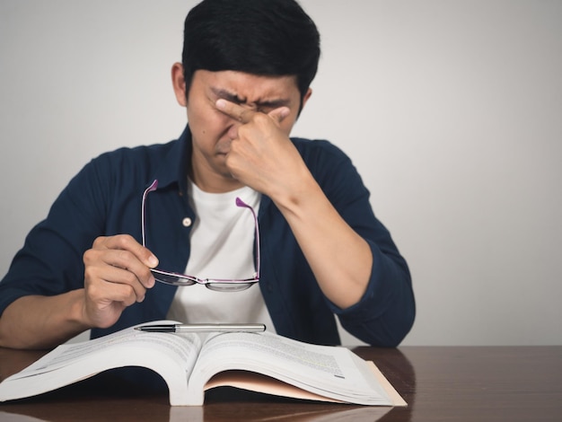 O homem asiático tira os óculos se sente tentado a aprender na mesa com o livro de texto