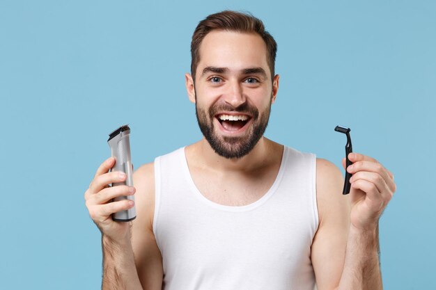 O homem barbudo de 20 anos de idade na camisa branca segura a navalha plástica elétrica escolhida isolada no retrato azul do estúdio do fundo pastel. Conceito de procedimentos cosméticos de saúde de cuidados com a pele. Simule o espaço da cópia.