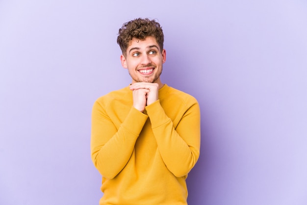 O homem caucasiano do cabelo encaracolado louro novo isolado mantém as mãos sob o queixo, está olhando felizmente de lado.