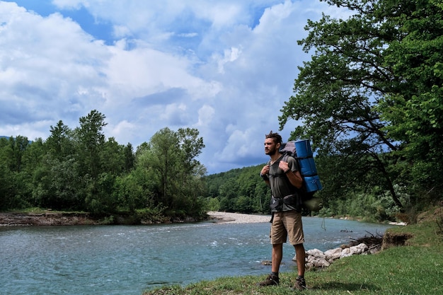 O homem caucasiano está caminhando ao longo da margem do rio da montanha com uma grande mochila de caminhada e desfrutando