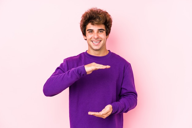 O homem caucasiano novo contra uma parede cor-de-rosa isolou guardar algo com ambas as mãos, apresentação do produto.