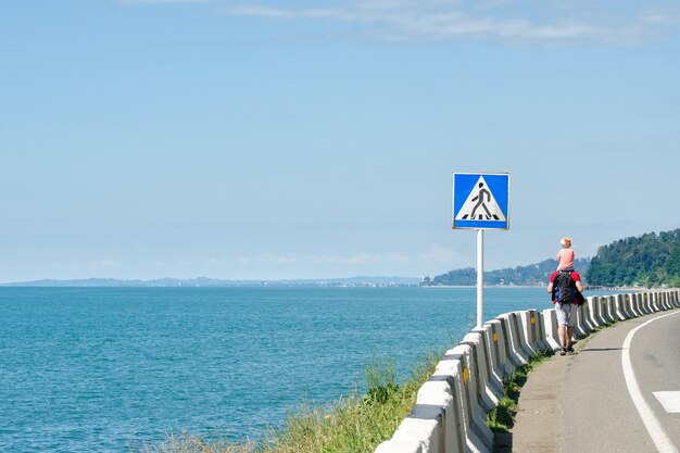 O homem com seu filho nos ombros caminha ao longo da estrada contra a parede do mar. sinal de travessia de pedestres