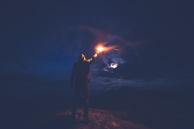 O homem com uma vara de fogo de artifício parado nas montanhas à noite