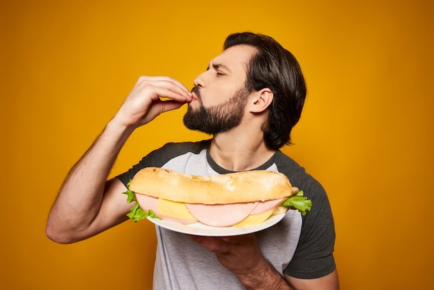 O homem considerável com sanduíche grande faz o gesto delicioso.