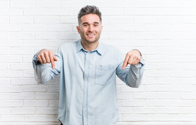 O homem considerável novo contra uma parede de tijolos aponta para baixo com os dedos, sentimento positivo.