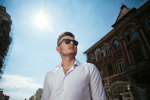 O homem da moda em uma camisa branca está parado na rua em um dia ensolarado está posando assistindo segurando óculos