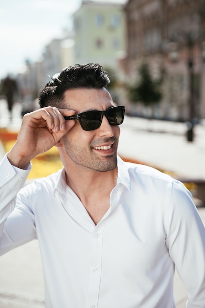 O homem da moda em uma camisa branca está parado na rua em um dia ensolarado está posando assistindo segurando óculos