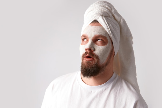 O homem de camiseta branca aplica o suporte de máscara facial contra um fundo branco. Tratamento de beleza, aliciamento e conceito de bem-estar. Procedimento de spa matinal de autocuidado