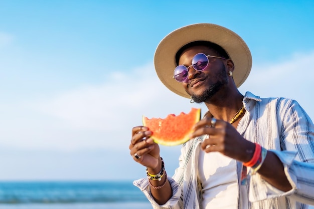 O homem de etnia negra aproveita as férias de verão na praia comendo uma melancia com um guarda-sol e óculos de sol