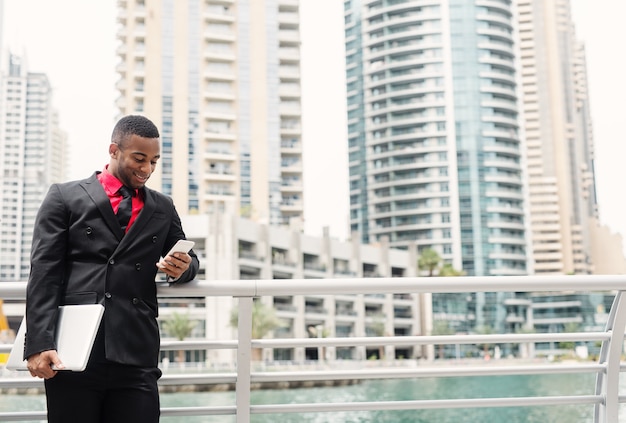 O homem de negócio afro-americano moderno novo inclinou-se na cerca no fuzileiro naval de Dubai ao olhar seu telefone com sorriso.