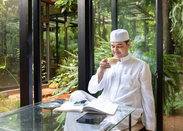 O homem de negócio muçulmano envelhecido asiático meados de senta-se no café bebe o café com telefone móvel esperto na tabela.