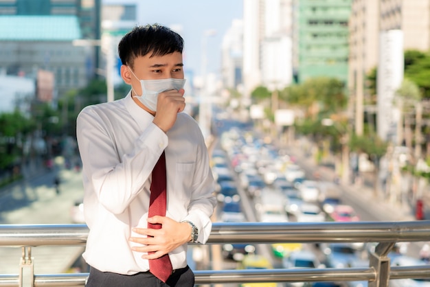 O homem de negócios asiático novo na camisa branca que vai trabalhar sentindo-se doente com tosse veste a máscara de proteção impede a poeira PM2.5, a poluição atmosférica, a poluição do ar e o COVID-19 com engarrafamento em Bangkok, Tailândia.