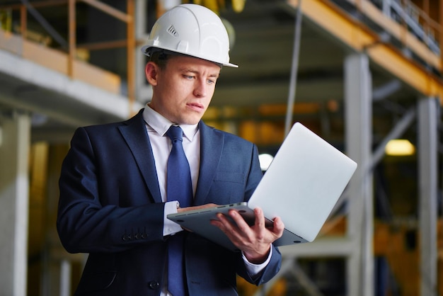 O homem de terno e capacete segura o computador portátil e olha para ele em um prédio industrial