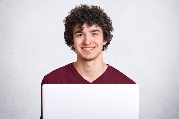 O homem deleitado positivo com o cabelo torrado, vestido na camisa ocasional de t, tem um sorriso encantador, usa o laptop moderno procurando a Internet, isolada no branco. Conceito de pessoas e tecnologia