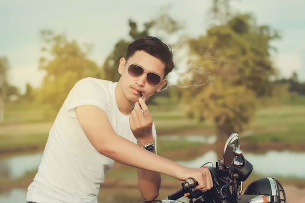 O homem do motociclista que senta-se fuma com seu velomotor ao lado do lago natural e bonito.