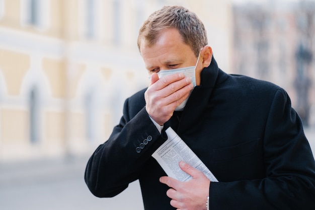 O homem doente tosse, cobre a boca com a palma da mão, usa máscara médica, tem sintomas de alergia, gripe, gripe ou coronavírus, caminha ao ar livre, segura jornal, sente-se mal. Covid-19, conceito de quarentena