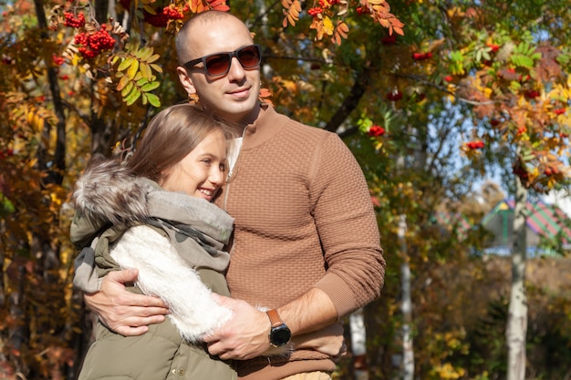 O homem dos óculos de sol abraça a filha bonita no fundo de árvores coloridas do outono