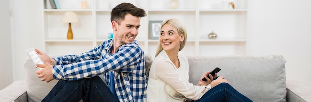 O homem e a mulher felizes com um telefone e um tablet sentam-se no sofá