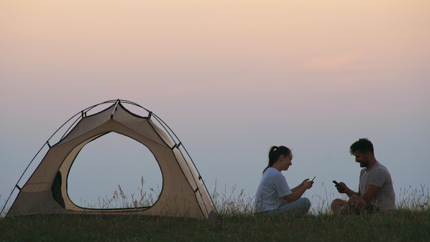 O homem e a mulher sentados perto da barraca do acampamento com smartphones