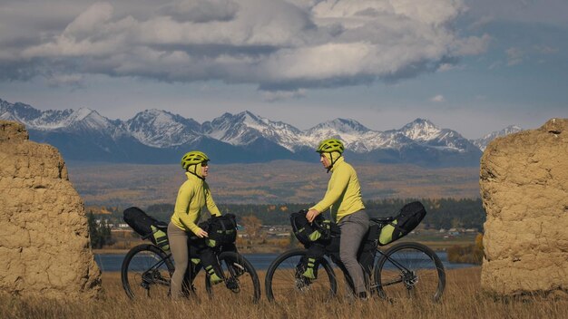 O homem e a mulher viajam em terreno misto cicloturismo com bikepacking As duas pessoas viajam com bolsas de bicicleta Roupa esportiva esportiva em cores pretas verdes Arco de pedra coberto de neve de montanha