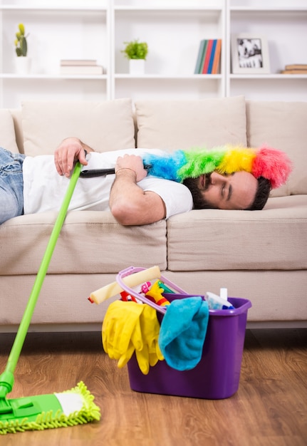 O homem é cansado e dormindo no sofá durante a limpeza em casa.