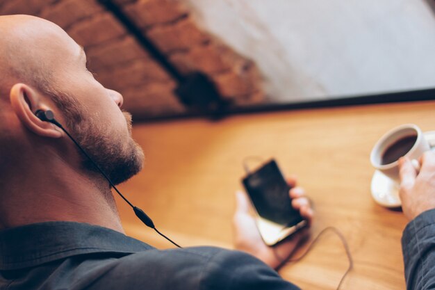 O homem em fones de ouvido usando telefone celular, sentado com café no café