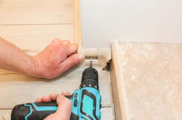 O homem está instalando o rodapé com uma furadeira. O reparo funciona dentro de casa. Reforma no apartamento.