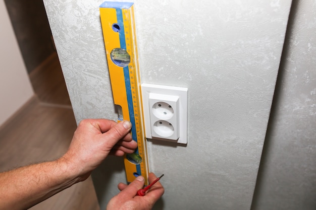 O homem está instalando o soquete. Nível de espírito nas mãos. Obras de reparação de manutenção de eletricidade no apartamento. Restauração dentro de casa.