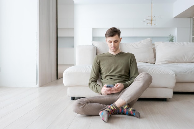 O homem está sentado no chão com os pés erguidos e olhando para o telemóvel.