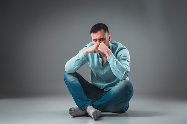 O homem está sentado no chão, isolado em um fundo cinza. Homem mostrando emoções diferentes.