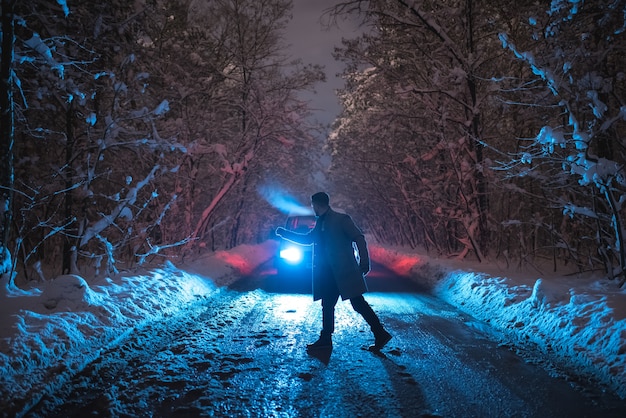 O homem fica na estrada de neve contra o carro. noite noite