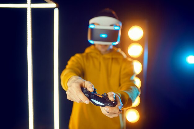 O homem joga o jogo usando capacete de realidade virtual e gamepad em cubo luminoso, vista frontal. Interior escuro do clube, tecnologia VR com visão 3D