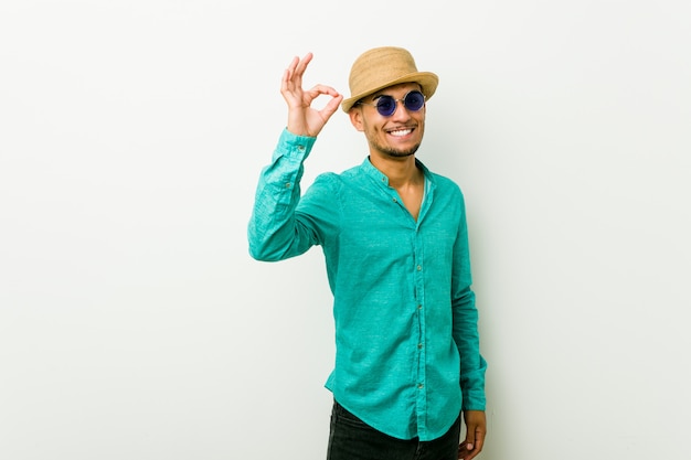 Foto o homem latino-americano novo que veste um verão veste alegre e seguro mostrando o gesto aprovado.