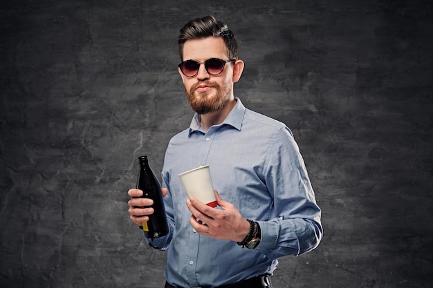 O homem moderno barbudo elegante segura uma xícara de café de papel e cerveja artesanal engarrafada.