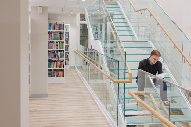 O homem na biblioteca está lendo o livro sentado nos degraus da escada de vidro