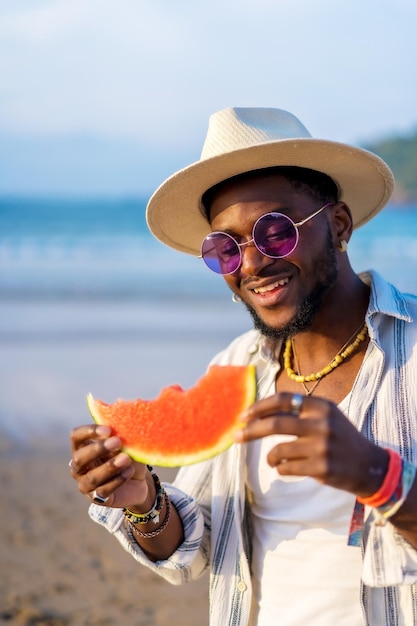 O homem negro étnico aproveita as férias de verão na praia comendo uma melancia sorrindo