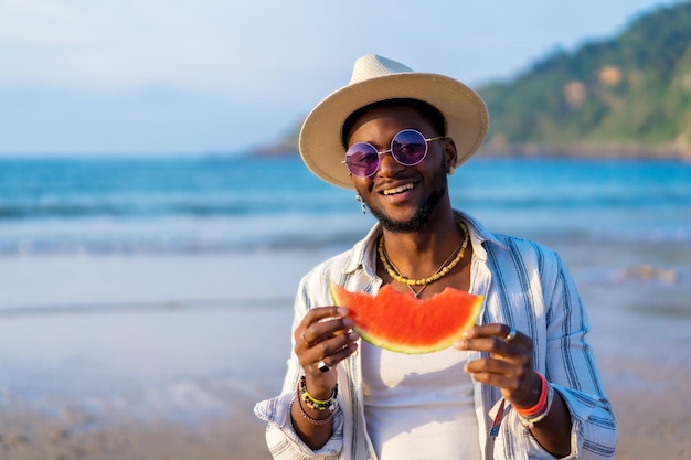 O homem negro étnico aproveita as férias de verão na praia comendo uma melancia