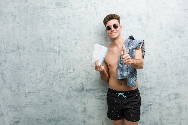 O homem novo que veste um roupa de banho que guarda passagens aéreas com polegares levanta, felicidades sobre algo, apoia e respeita o conceito.