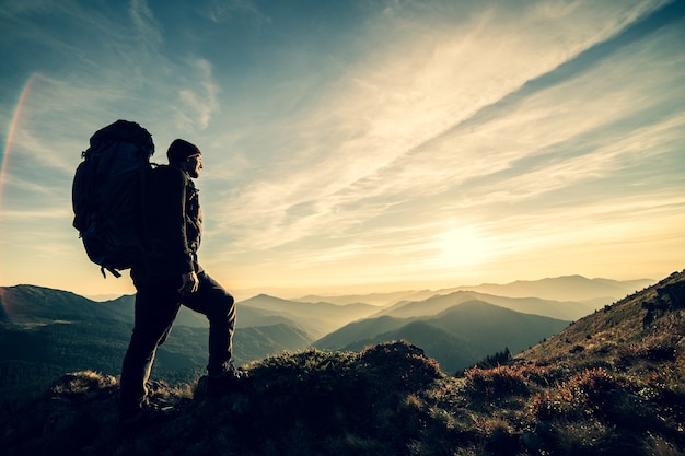 O homem parado com uma mochila de camping na rocha com um pôr do sol pitoresco