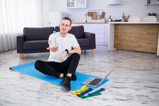 O homem pratica esportes em casa. Atleta bebe água sentado no chão em casa, estilo de vida saudável