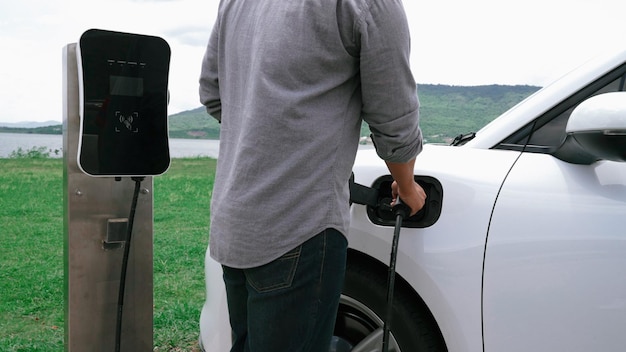 O homem progressivo conecta o plugue do cabo de alimentação da estação de carregamento ao seu carro elétrico EV movido por energia verde em campo verde para o futuro conceito de ambiente limpo