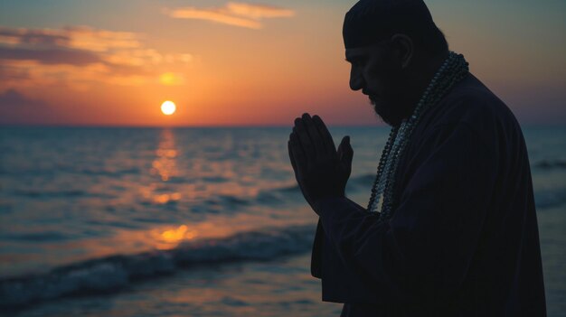 O homem segura contas de oração e reza ao pôr-do-sol com o oceano tranquilo como pano de fundo.