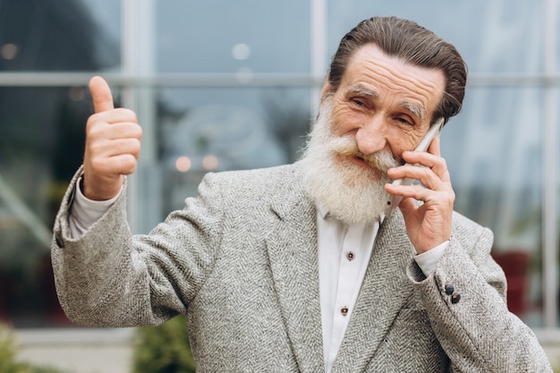 O homem sênior com uma barba grisalha e bigode em uma jaqueta fala ao telefone