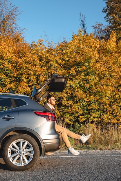O homem senta-se no porta-bagagens do seu carro e desfruta do sol de outono enquanto ele brilha nele
