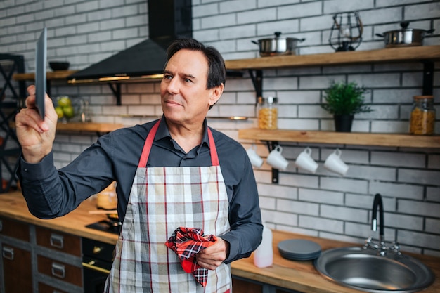 O homem sério no avental fica na cozinha e olha o prato na mão. Ele secou. Também cara segurar toalha de cozinha.