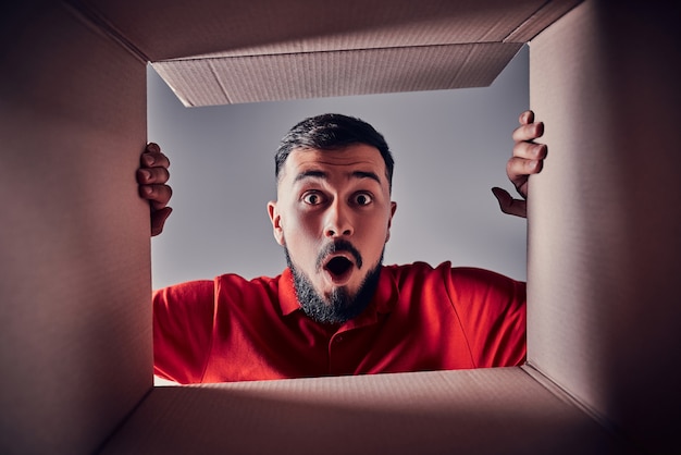 Foto o homem surpreso abrindo a caixa de papelão e olhando para dentro. o pacote, entrega, surpresa, presente, conceito de estilo de vida. conceitos de emoções humanas e expressões faciais