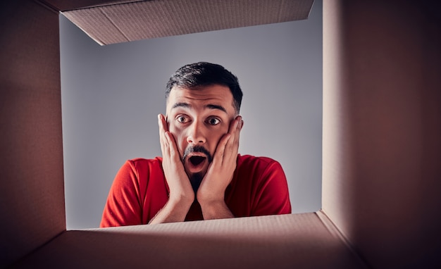 O homem surpreso abrindo a caixa de papelão e olhando para dentro. O pacote, entrega, surpresa, presente, conceito de estilo de vida. Conceitos de emoções humanas e expressões faciais